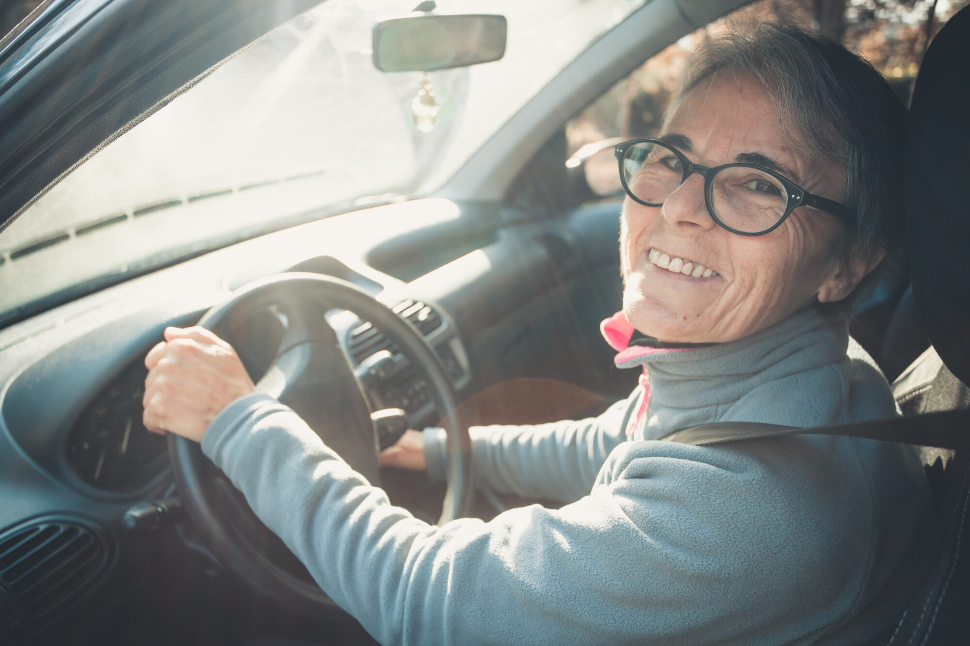 An elderly woman in a pair of black glasses and a blue fleece smiles at the camera from inside her car
